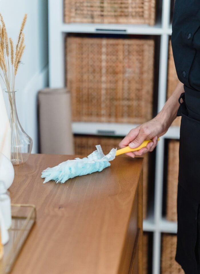 Person dusting a wooden cabinet with a feather duster in a bright, modern home.