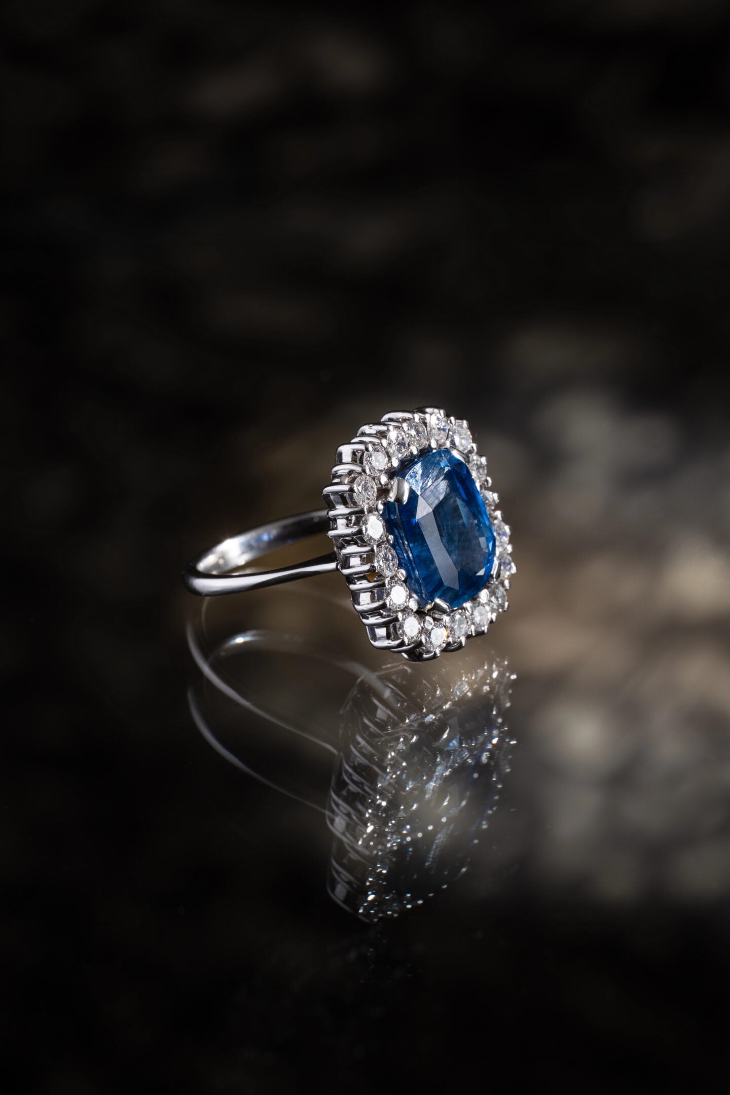 White gold ring featuring a deep blue cushion-cut sapphire surrounded by a halo of sparkling diamonds, displayed on a reflective dark surface.
