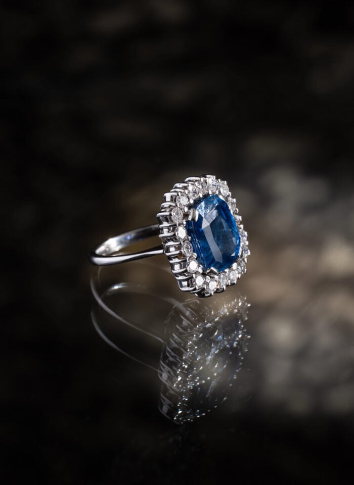 White gold ring featuring a deep blue cushion-cut sapphire surrounded by a halo of sparkling diamonds, displayed on a reflective dark surface.