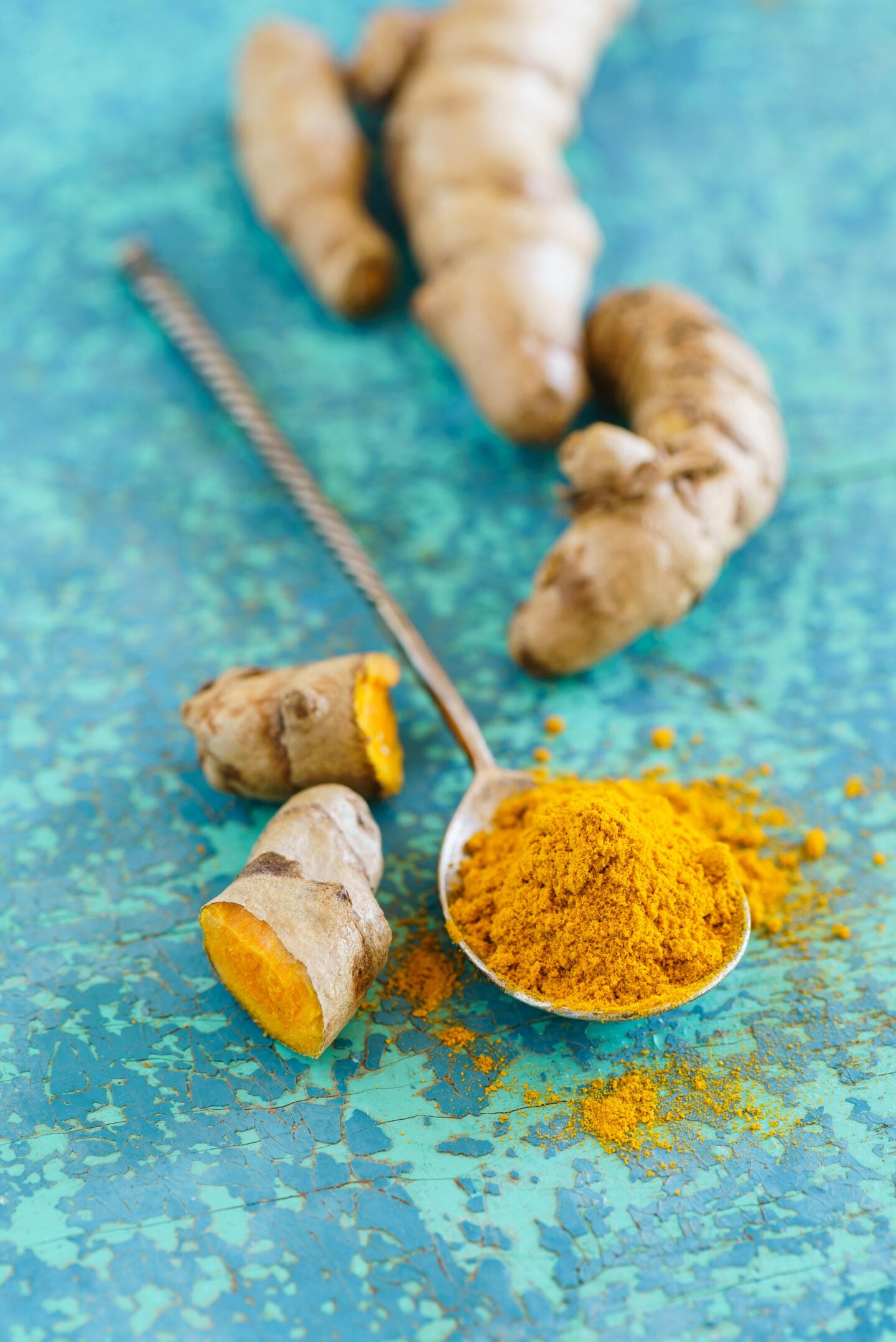 Fresh turmeric root and ground turmeric powder in a spoon on a blue surface.