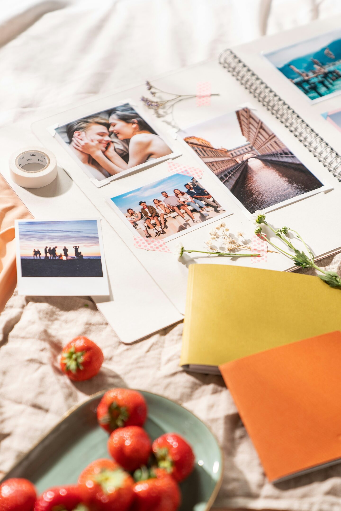 Open scrapbook filled with printed photographs of couples, friends and travel memories, decorated with dried flowers and washi tape, alongside photo prints, colourful notebooks and a bowl of strawberries.