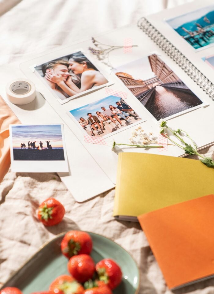 Open scrapbook filled with printed photographs of couples, friends and travel memories, decorated with dried flowers and washi tape, alongside photo prints, colourful notebooks and a bowl of strawberries.