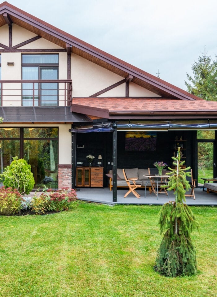 Modern two storey house with a covered outdoor living area, patio furniture and a large landscaped garden surrounded by trees.