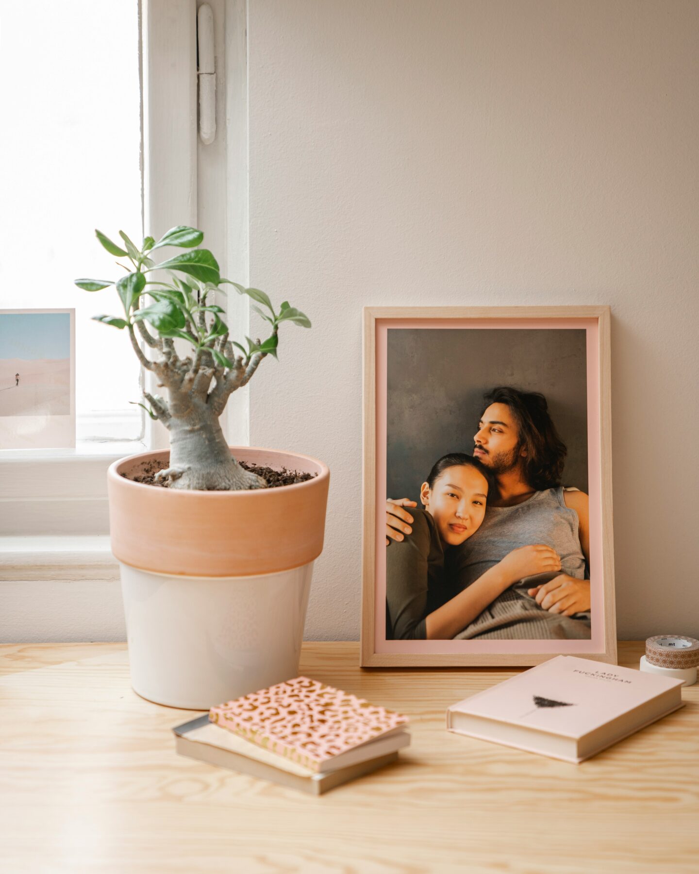 Framed printed photograph of a couple embracing displayed on a wooden table beside a potted plant and small notebooks in a bright, cosy home interior.