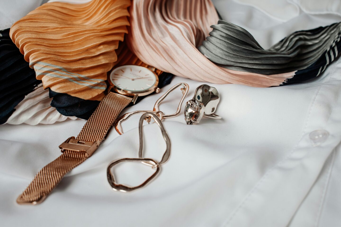 Gold watch and statement earrings arranged on a white shirt beside a pleated scarf in warm orange, blush pink, grey and black tones.