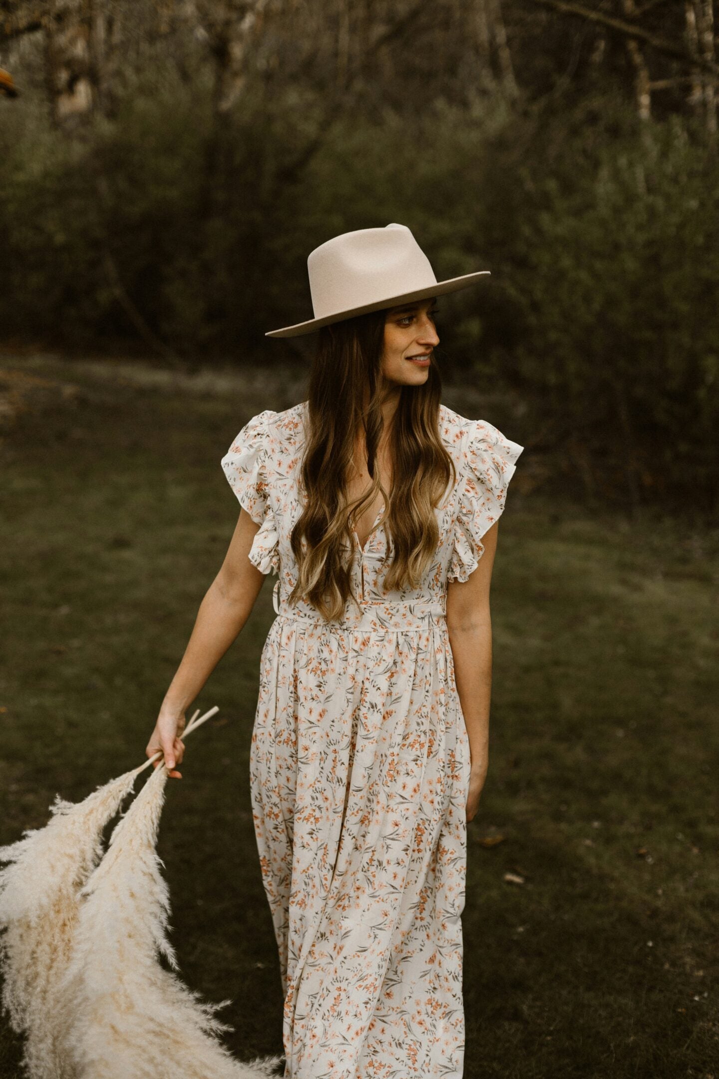 Maxi dresses - woman wearing a flowing white floral maxi dress and beige wide-brim hat outdoors, holding dried pampas grass against a woodland backdrop.