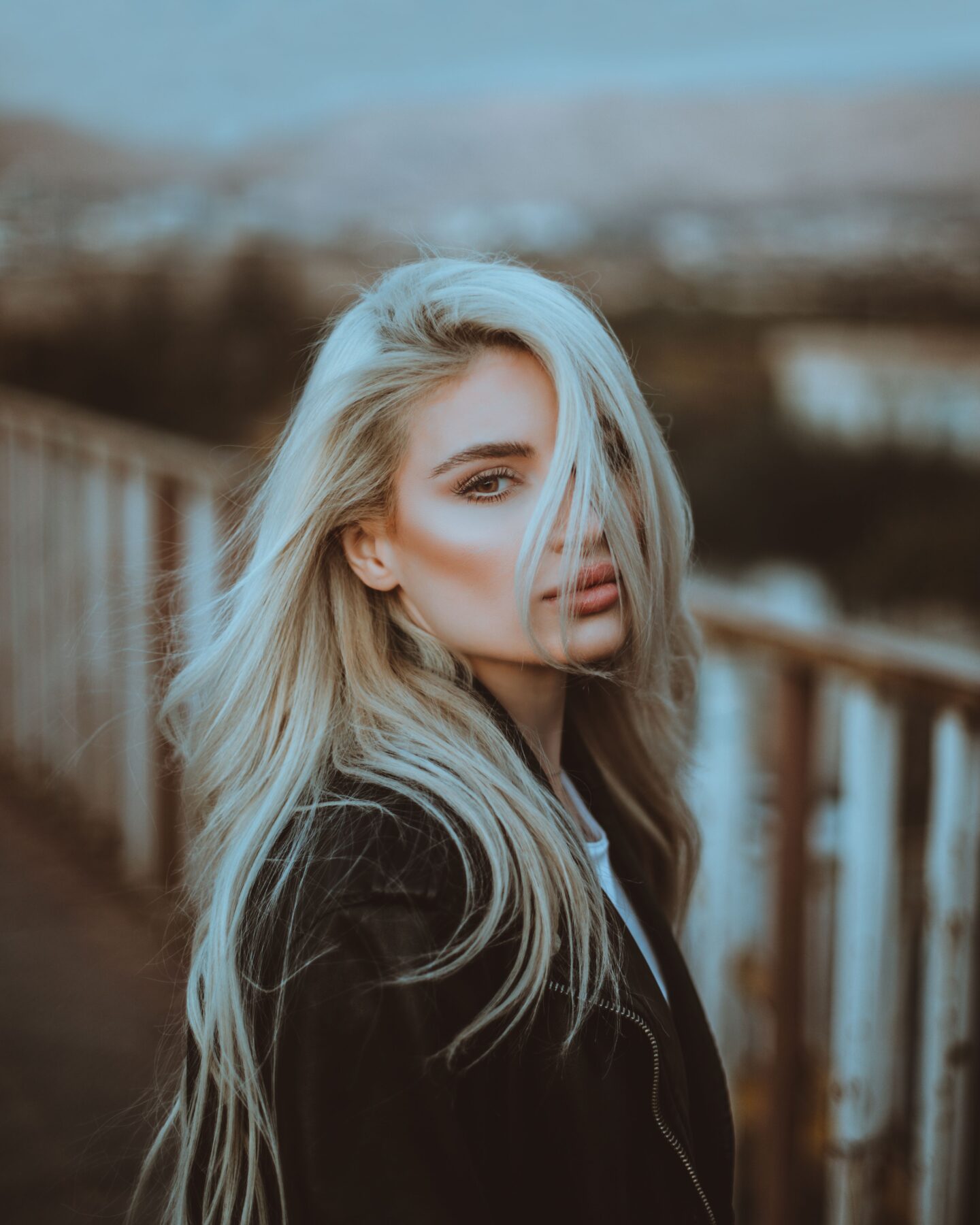 Close-up portrait of a woman with long, voluminous, tousled platinum blonde hair cascading over her shoulders. Loose strands fall across her face, partially covering one eye. She has defined eyebrows, long eyelashes, and nude pink lips. She is wearing a black leather jacket and standing outdoors beside a wooden railing, with a softly blurred background.