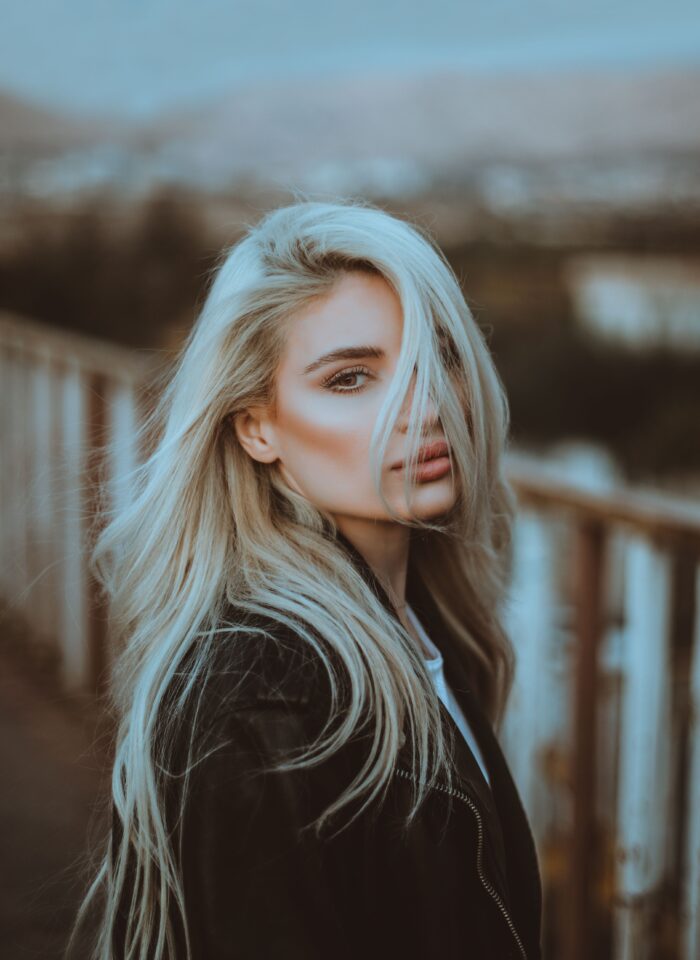 Close-up portrait of a woman with long, voluminous, tousled platinum blonde hair cascading over her shoulders. Loose strands fall across her face, partially covering one eye. She has defined eyebrows, long eyelashes, and nude pink lips. She is wearing a black leather jacket and standing outdoors beside a wooden railing, with a softly blurred background.