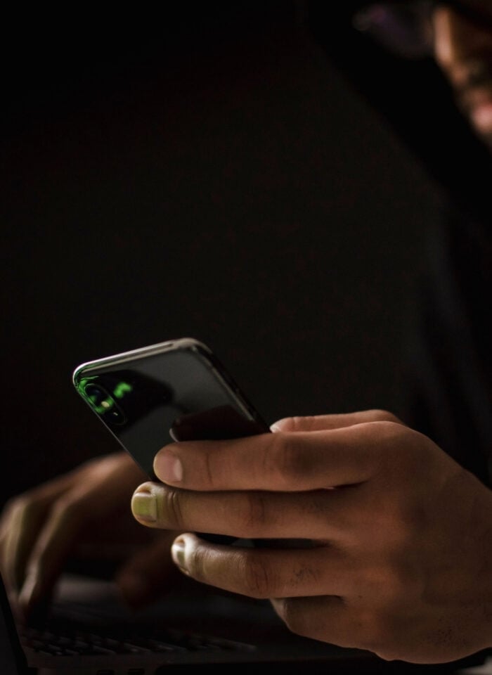A hacker wearing dark clothing sits in a dimly lit room, using a smartphone while working on a laptop. The low-key lighting obscures the person’s face and creates a dark, secretive atmosphere associated with cybersecurity, hacking, online threats, and cybercrime.