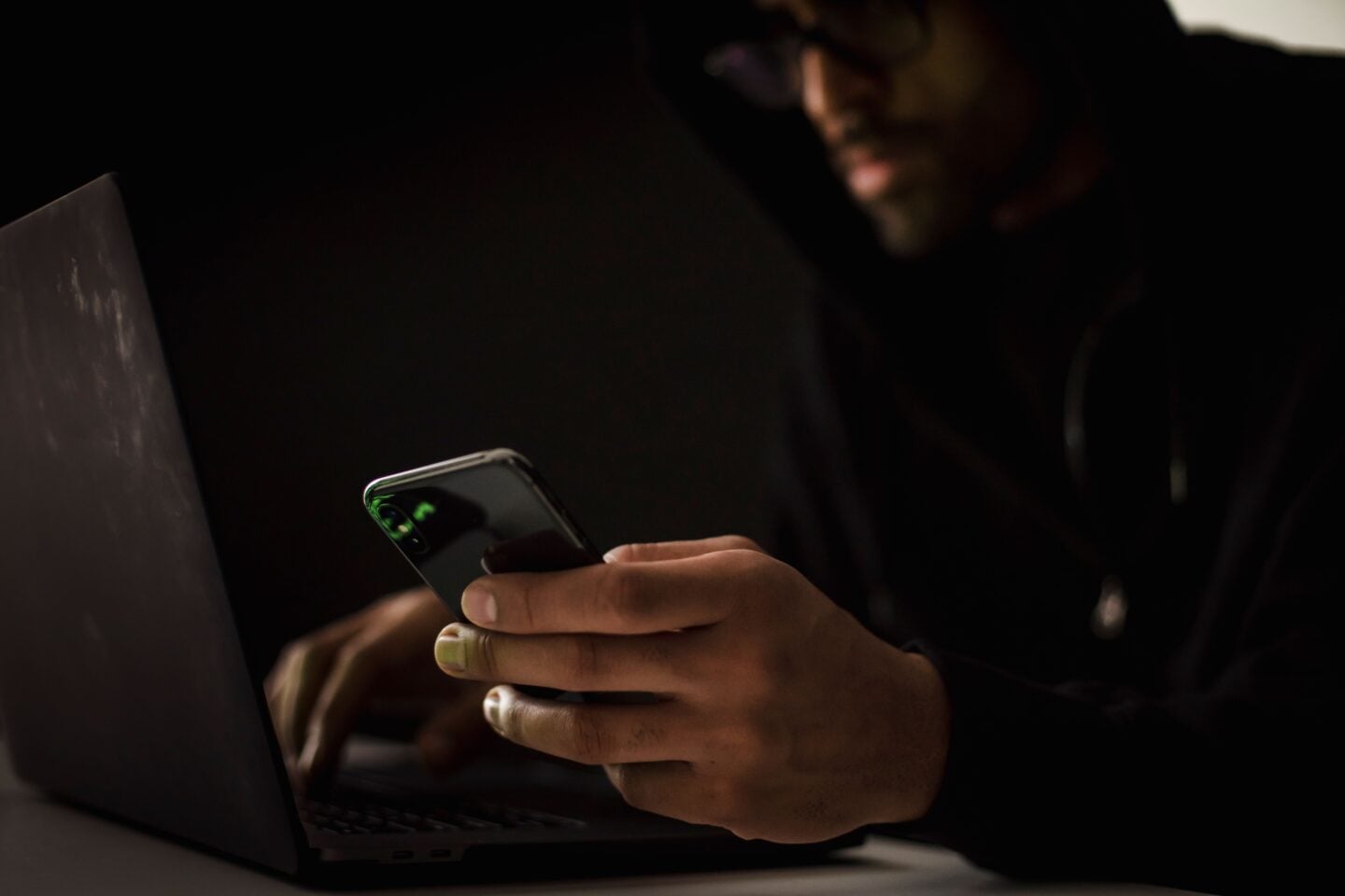 A hacker wearing dark clothing sits in a dimly lit room, using a smartphone while working on a laptop. The low-key lighting obscures the person’s face and creates a dark, secretive atmosphere associated with cybersecurity, hacking, online threats, and cybercrime.