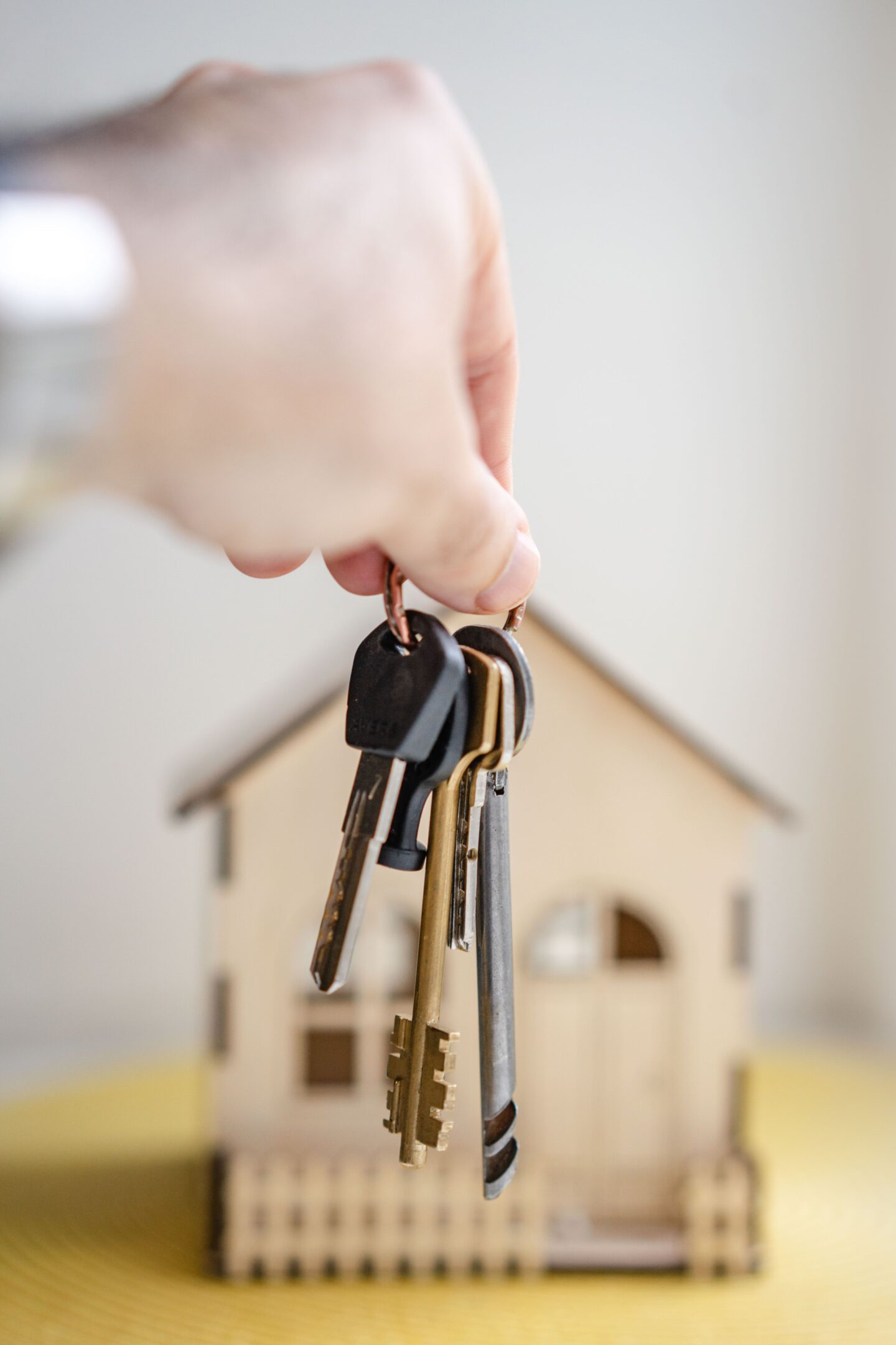 Close-up of a hand holding a set of metal house keys on a keyring in front of a small wooden model house with a pitched roof, windows, and a front porch, against a softly blurred light background.