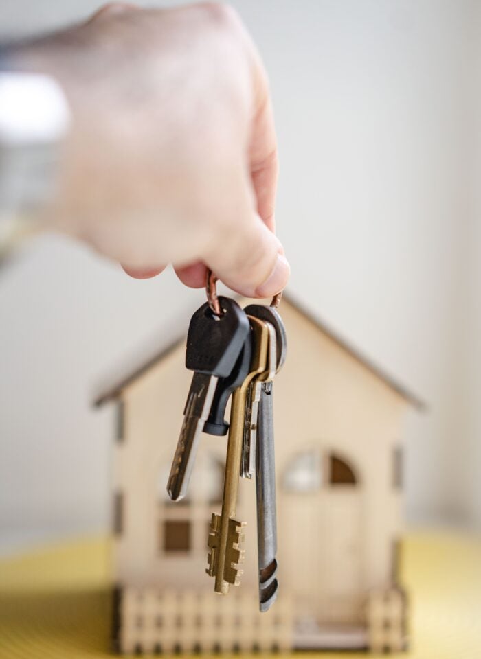 Close-up of a hand holding a set of metal house keys on a keyring in front of a small wooden model house with a pitched roof, windows, and a front porch, against a softly blurred light background.