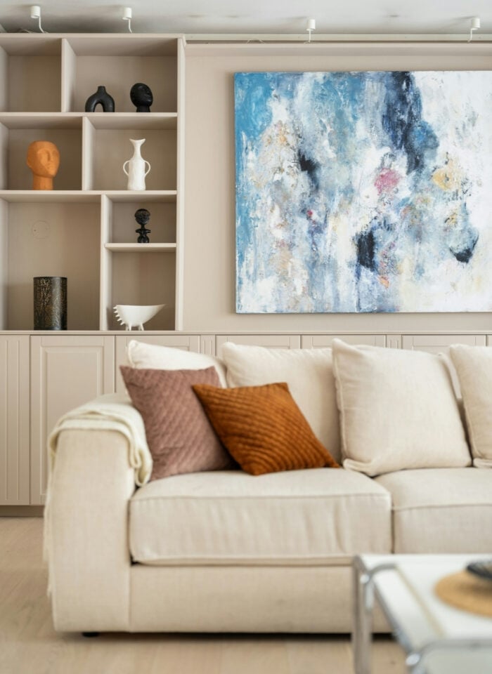 Neutral living room with a cream sofa, rust and blush pillows, built-in display shelves, and a large blue-and-white abstract painting.