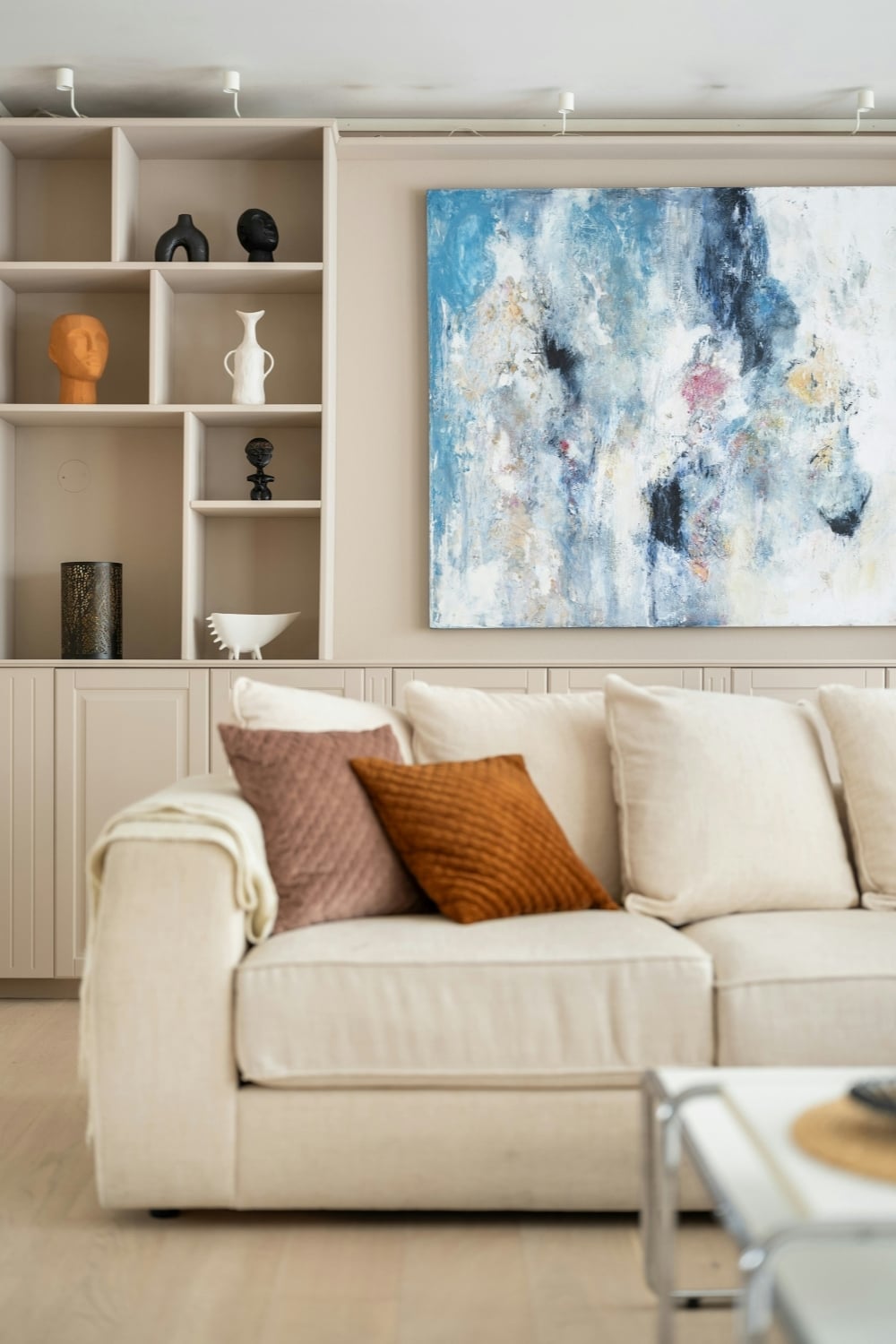 Neutral living room with a cream sofa, rust and blush pillows, built-in display shelves, and a large blue-and-white abstract painting.