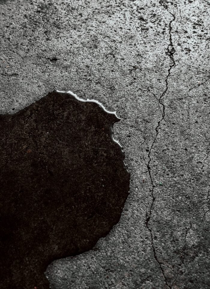 Close-up of a rough, grey concrete floor with a large, irregularly shaped puddle of dark water spreading across the surface. A thin, jagged crack runs vertically through the concrete beside the puddle, with smaller hairline fractures and tiny pits visible in the textured flooring. The wet area contrasts sharply with the surrounding dry concrete, creating a dark, uneven outline with a slightly reflective edge. The image illustrates water pooling on a cracked concrete floor, a possible warning sign of an underground plumbing leak or slab leak beneath a home's foundation.