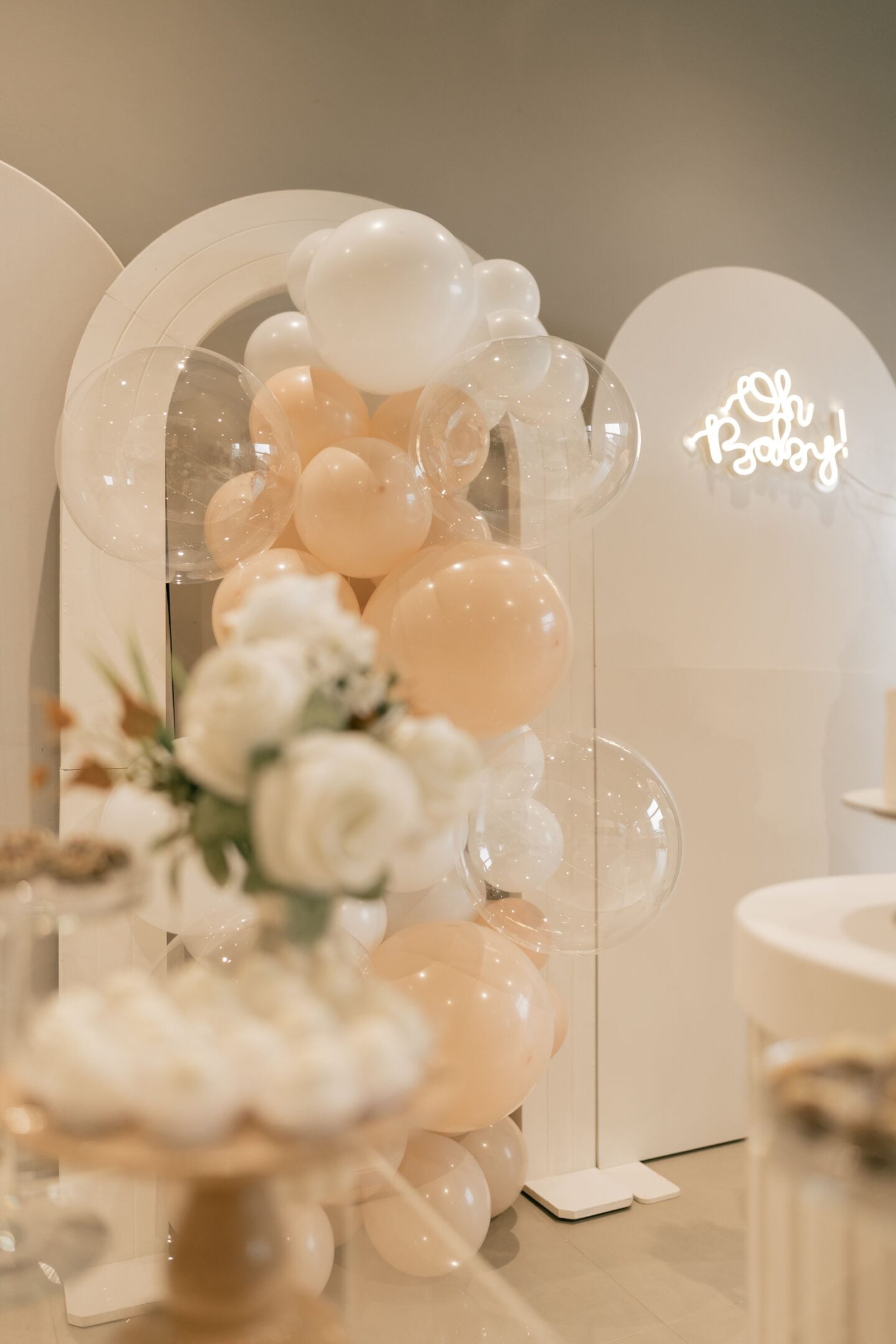 Elegant baby shower decorations featuring peach, white and transparent balloons, white floral arrangements and a glowing “Oh Baby!” sign.