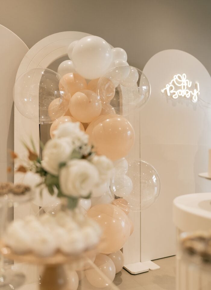 Elegant baby shower decorations featuring peach, white and transparent balloons, white floral arrangements and a glowing “Oh Baby!” sign.
