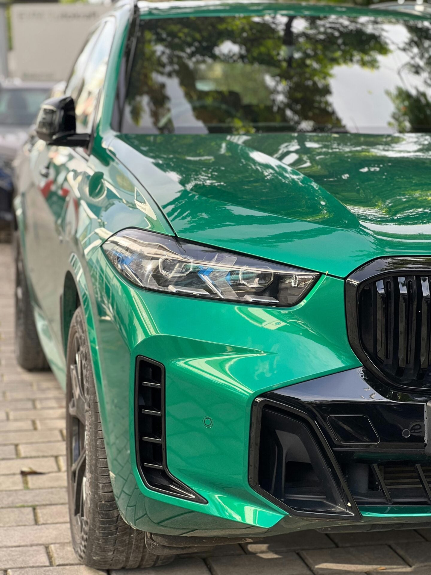 Close-up front three-quarter view of a bright green BMW X5 luxury SUV, showing its slim LED headlight, black kidney grille, sculpted hood, black front air intake and glossy green bodywork reflecting nearby trees.