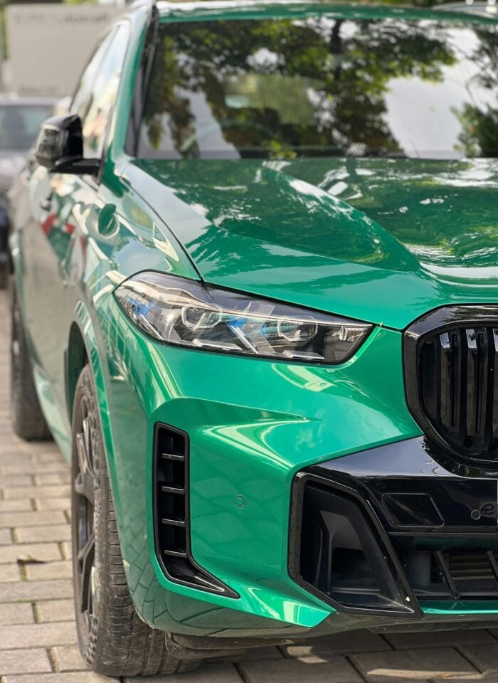 Close-up front three-quarter view of a bright green BMW X5 luxury SUV, showing its slim LED headlight, black kidney grille, sculpted hood, black front air intake and glossy green bodywork reflecting nearby trees.