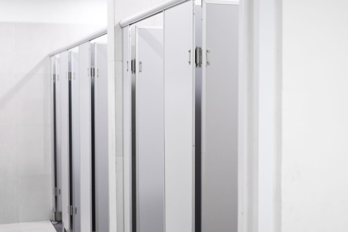 Row of white and light grey commercial restroom cubicle partitions with metal hinges, door handles, and overhead support rails in a clean, modern public bathroom with white tiled walls and a light grey tiled floor.