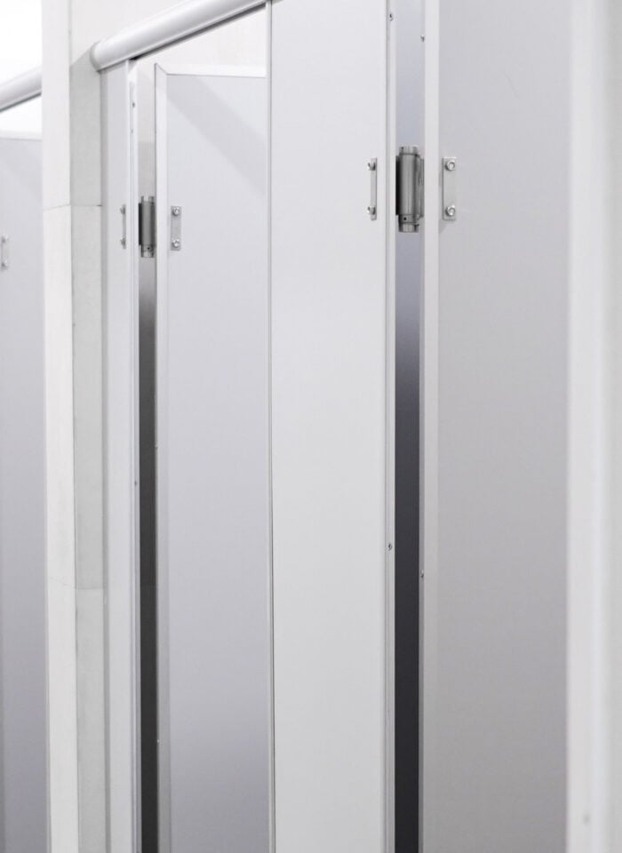 Row of white and light grey commercial restroom cubicle partitions with metal hinges, door handles, and overhead support rails in a clean, modern public bathroom with white tiled walls and a light grey tiled floor.