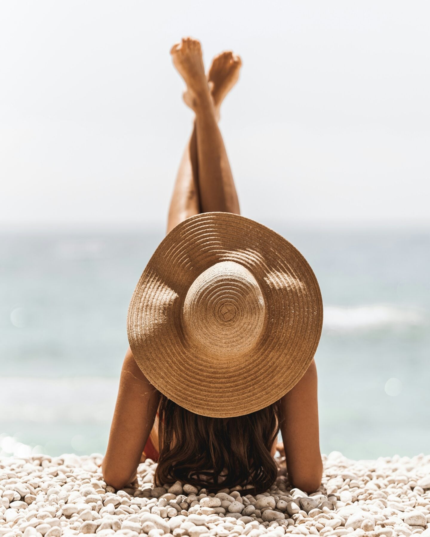 Woman wearing a wide-brimmed straw hat reclining on the white pebbles of Porto Katsiki beach in Greece, with her legs raised and turquoise sea behind her.