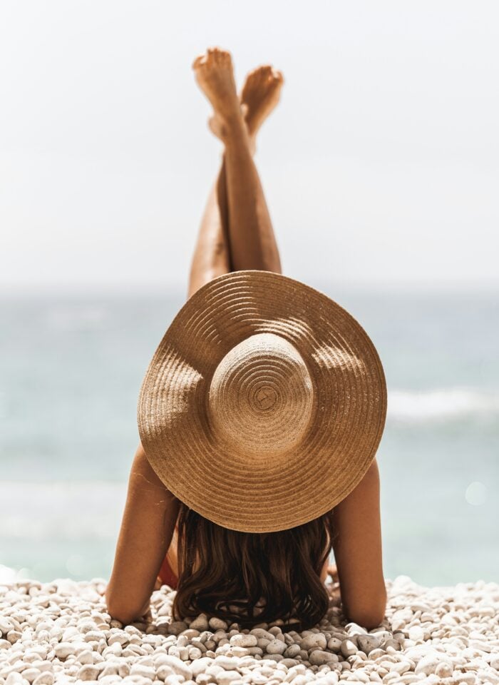 Woman wearing a wide-brimmed straw hat reclining on the white pebbles of Porto Katsiki beach in Greece, with her legs raised and turquoise sea behind her.