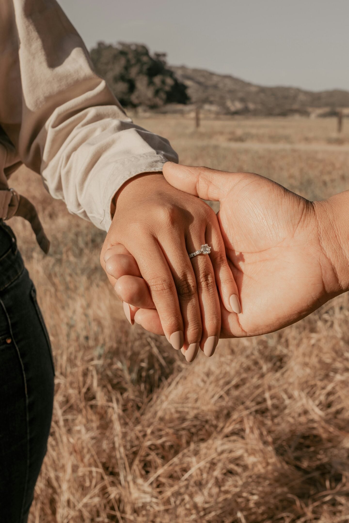 Close-up of a woman's hand wearing a delicate diamond engagement ring, gently held by her partner in a sunlit field of dry grass.