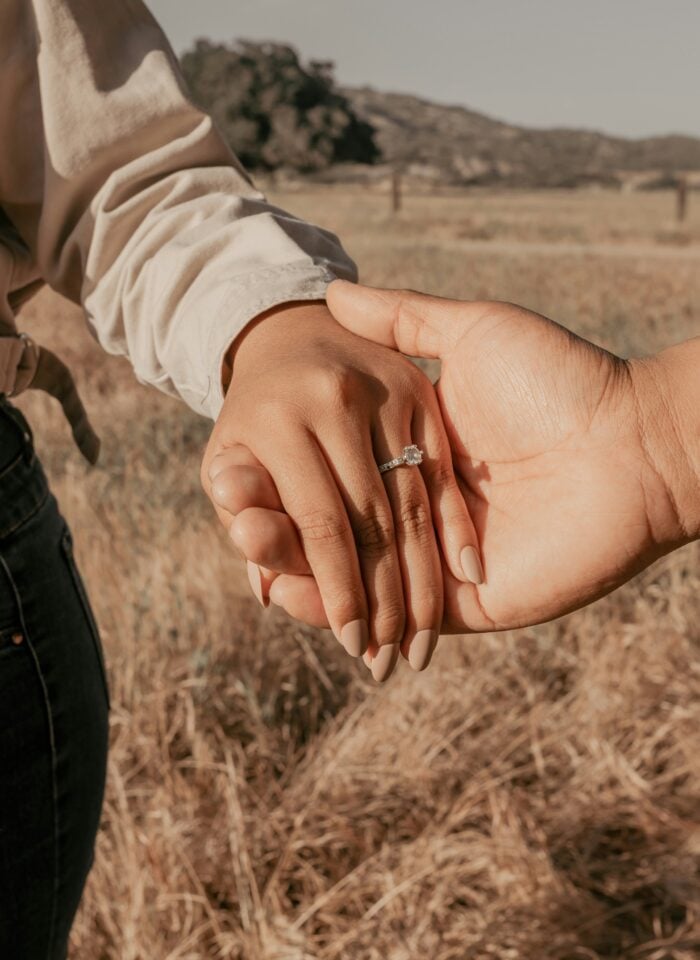 Close-up of a woman's hand wearing a delicate diamond engagement ring, gently held by her partner in a sunlit field of dry grass.