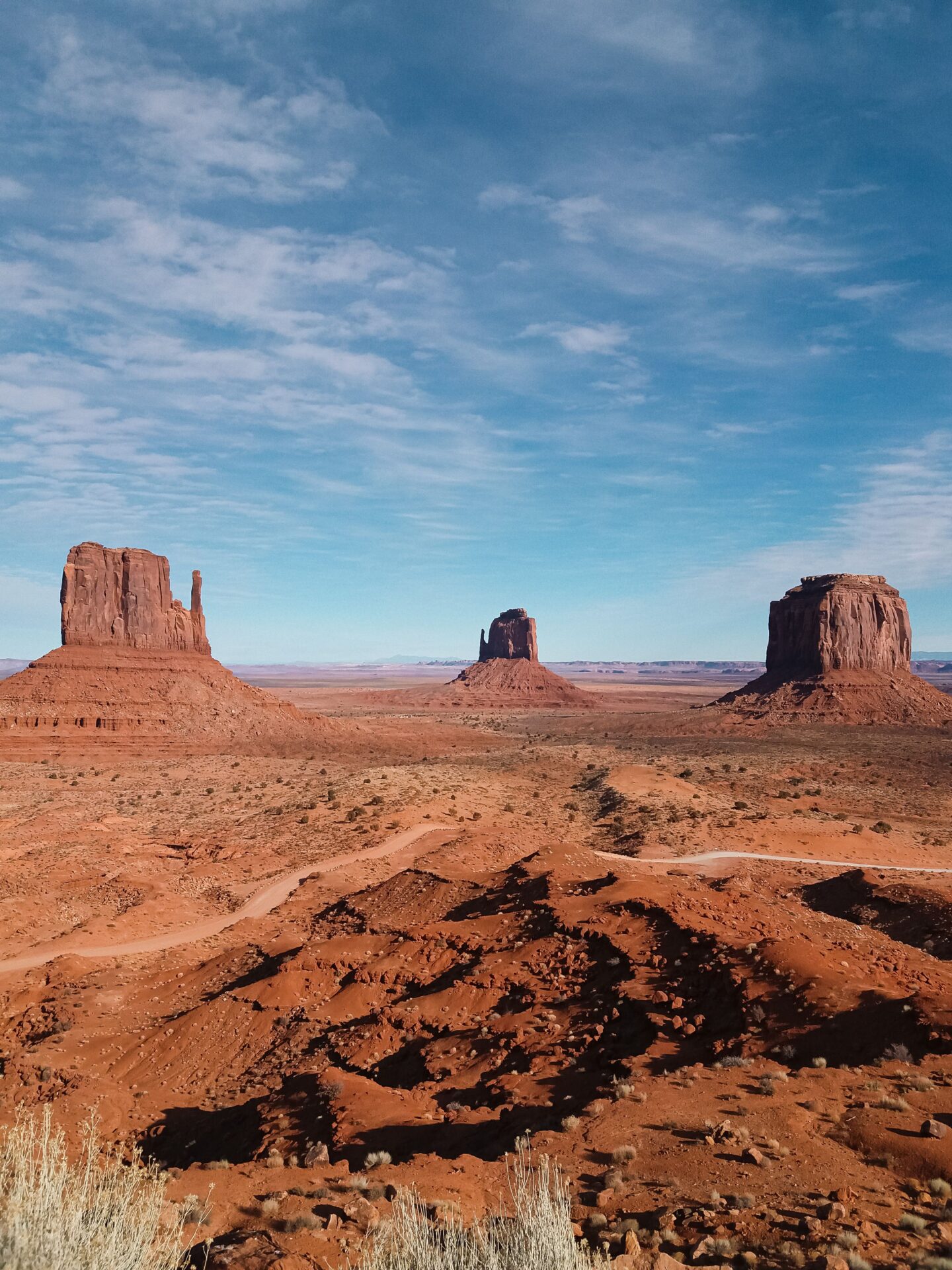 Panoramic view of Monument Valley on the Arizona–Utah border, featuring three iconic red sandstone rock formations known as West Mitten Butte, East Mitten Butte, and Merrick Butte rising dramatically from the vast desert landscape. The foreground reveals rugged, rust-red rocky terrain dotted with sparse desert grasses and low shrubs, with a winding dirt road cutting through the valley. The towering sandstone buttes stand against an expansive blue sky filled with delicate, wispy white clouds, showcasing the breathtaking natural beauty of the American Southwest.