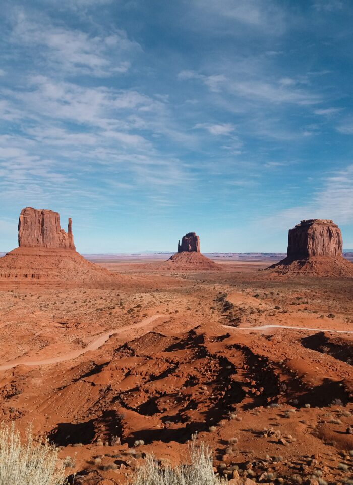 Panoramic view of Monument Valley on the Arizona–Utah border, featuring three iconic red sandstone rock formations known as West Mitten Butte, East Mitten Butte, and Merrick Butte rising dramatically from the vast desert landscape. The foreground reveals rugged, rust-red rocky terrain dotted with sparse desert grasses and low shrubs, with a winding dirt road cutting through the valley. The towering sandstone buttes stand against an expansive blue sky filled with delicate, wispy white clouds, showcasing the breathtaking natural beauty of the American Southwest.