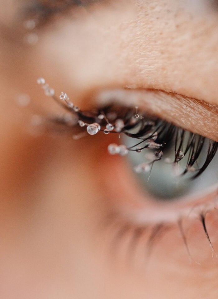 Extreme close-up of an eye with long, dark eyelashes covered in tiny water droplets, highlighting the delicate lashes and skin around the eyelid during cold winter weather.