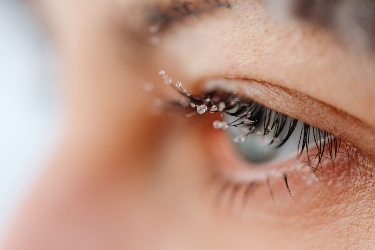 Extreme close-up of an eye with long, dark eyelashes covered in tiny water droplets, highlighting the delicate lashes and skin around the eyelid during cold winter weather.