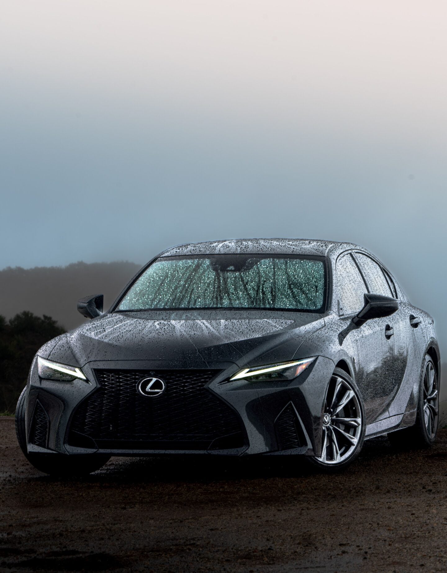 Dark gray Lexus IS luxury sports sedan photographed from the front in rainy weather, with water covering the bodywork and windshield, sharp LED headlights illuminated, a large black spindle grille and Lexus emblem prominently visible.