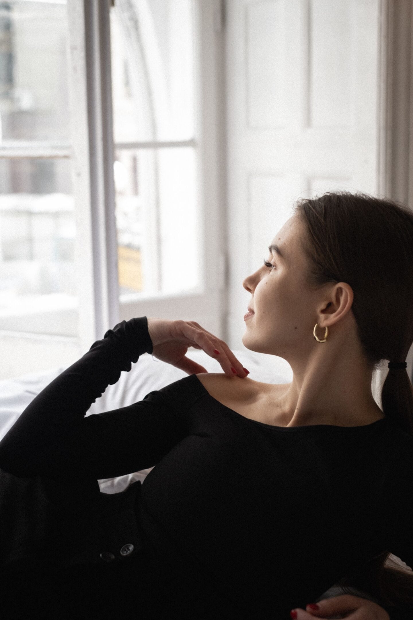 Woman wearing gold hoop earrings and a black off-the-shoulder top, sitting beside a window with her hair tied back, showcasing elegant minimalist jewelry and effortless winter fashion.