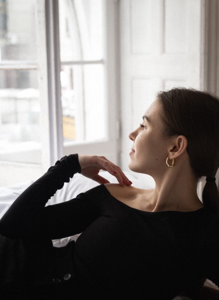 Woman wearing gold hoop earrings and a black off-the-shoulder top, sitting beside a window with her hair tied back, showcasing elegant minimalist jewelry and effortless winter fashion.