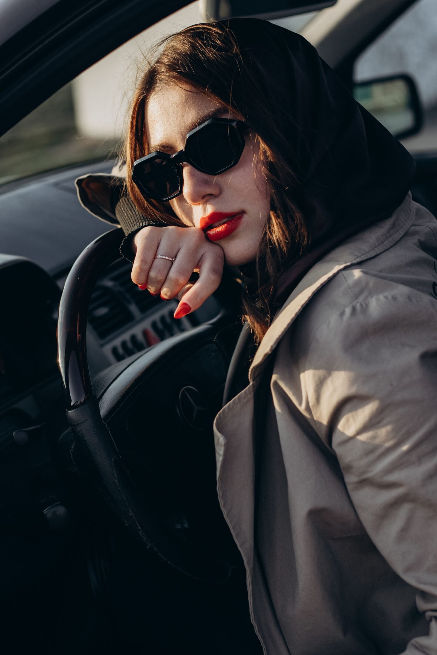Woman wearing black sunglasses and a beige coat sitting in a car’s driver’s seat, resting her hand on the steering wheel.