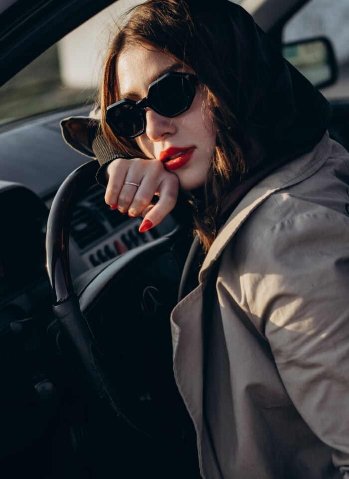 Woman wearing black sunglasses and a beige coat sitting in a car’s driver’s seat, resting her hand on the steering wheel.