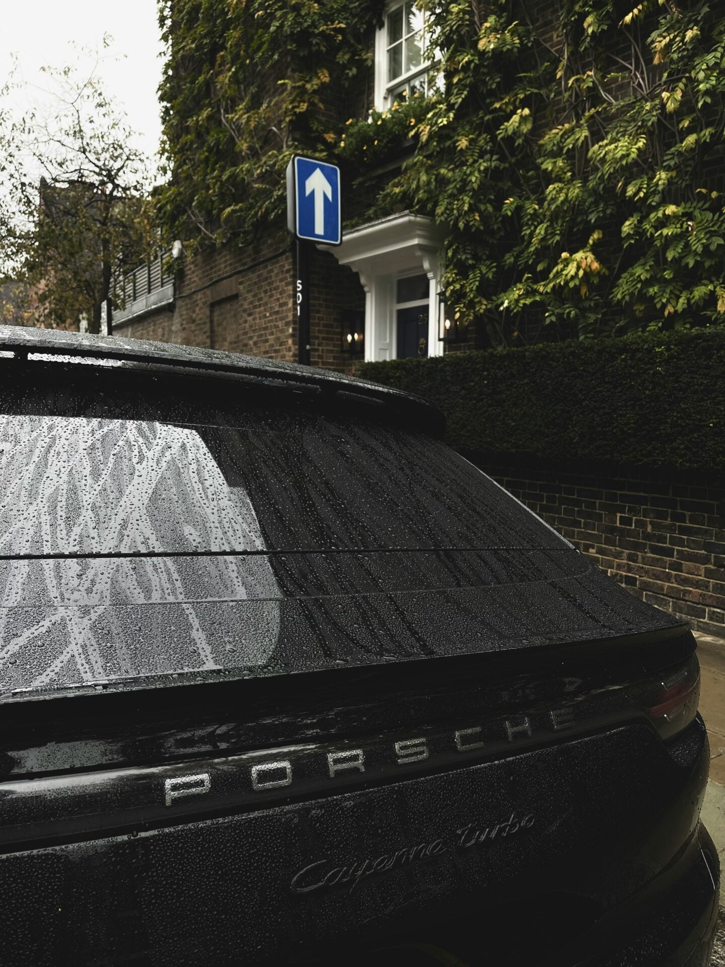 Rear close-up of a black Porsche Cayenne Turbo parked on a rain-soaked residential street, with water droplets covering the rear window and bodywork, Porsche lettering and Cayenne Turbo badge visible, and an ivy-covered brick house in the background.