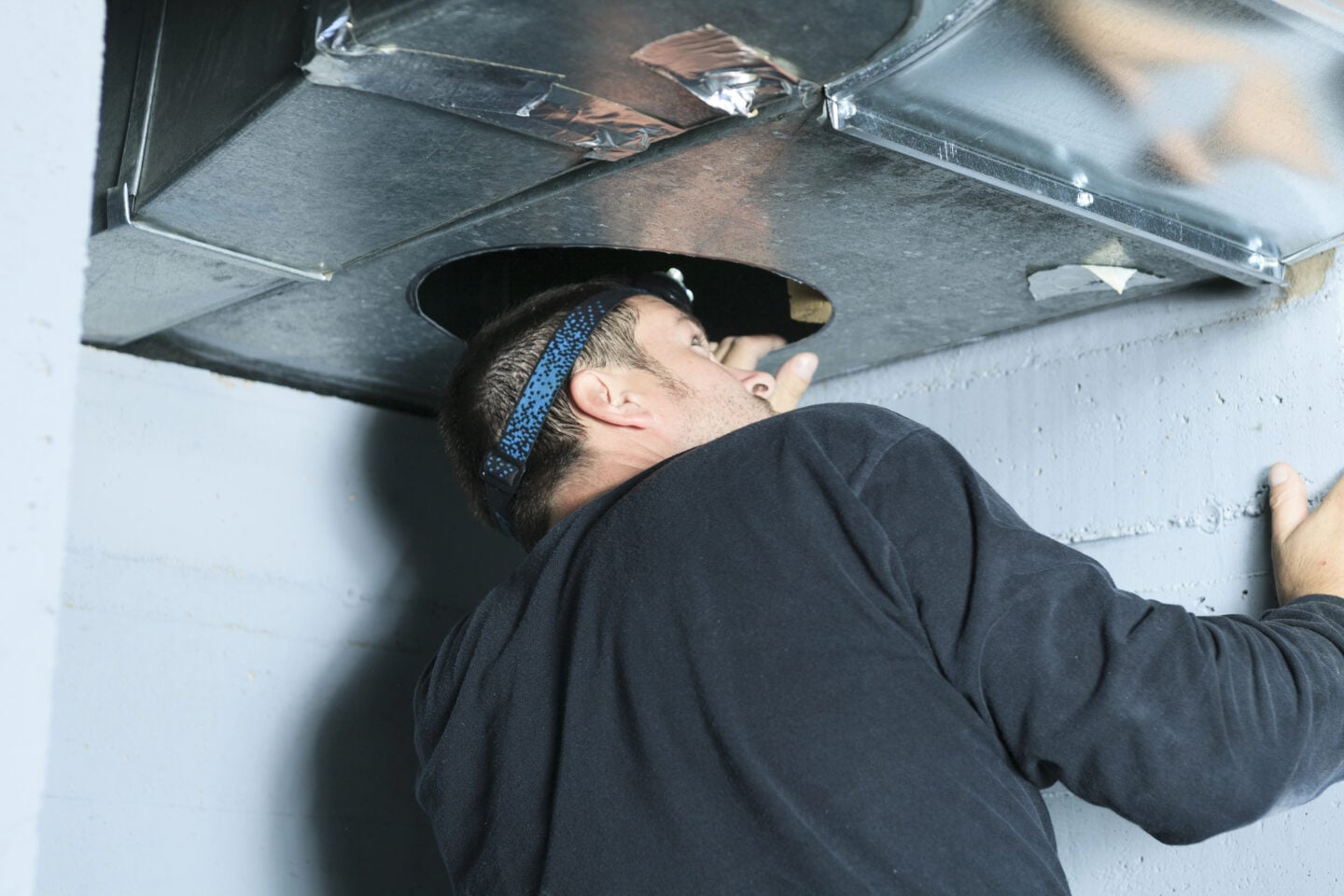 Technician wearing a headlamp inspects the inside of a metal air duct through a circular opening.