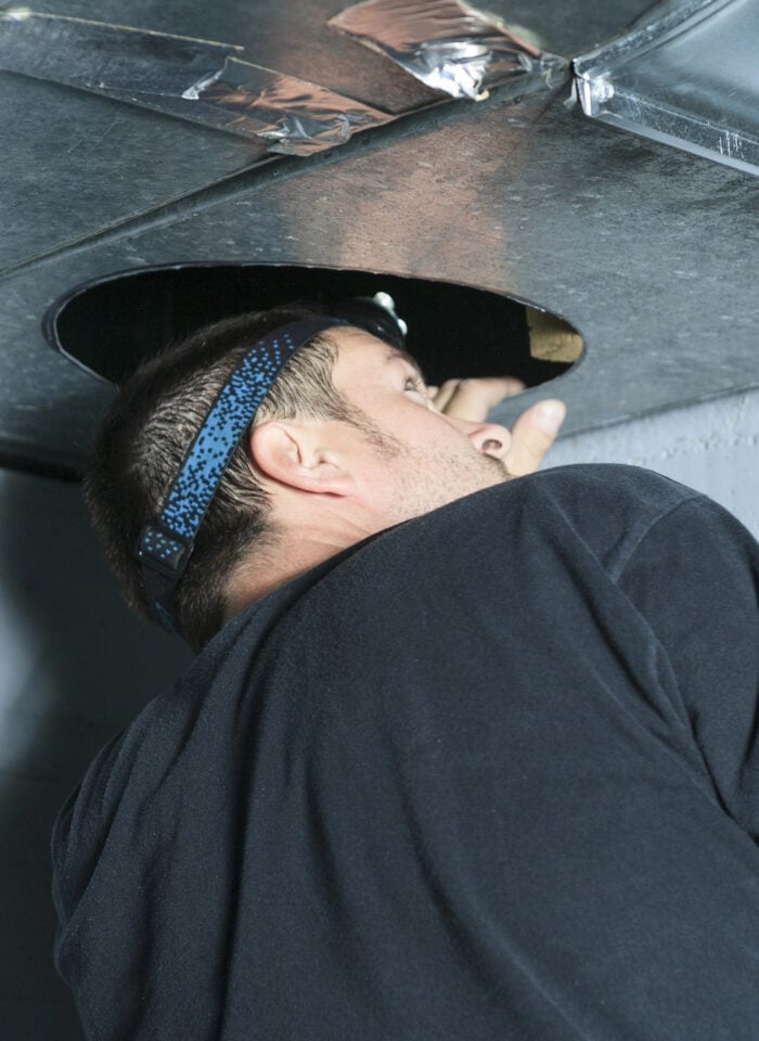 Technician wearing a headlamp inspects the inside of a metal air duct through a circular opening.