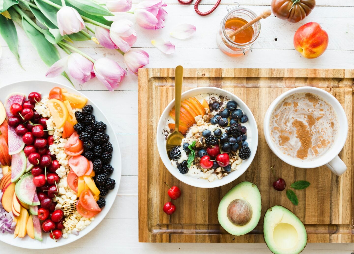 Overhead view of a colourful breakfast spread on a white wooden table. A bowl topped with blueberries, blackberries, cherries, sliced fruit, nuts and seeds sits on a wooden board beside a cup of coffee and two avocado halves. A large plate of assorted fruit and vegetables is on the left, with pink tulips, a jar of honey and additional fruit arranged around the table.
