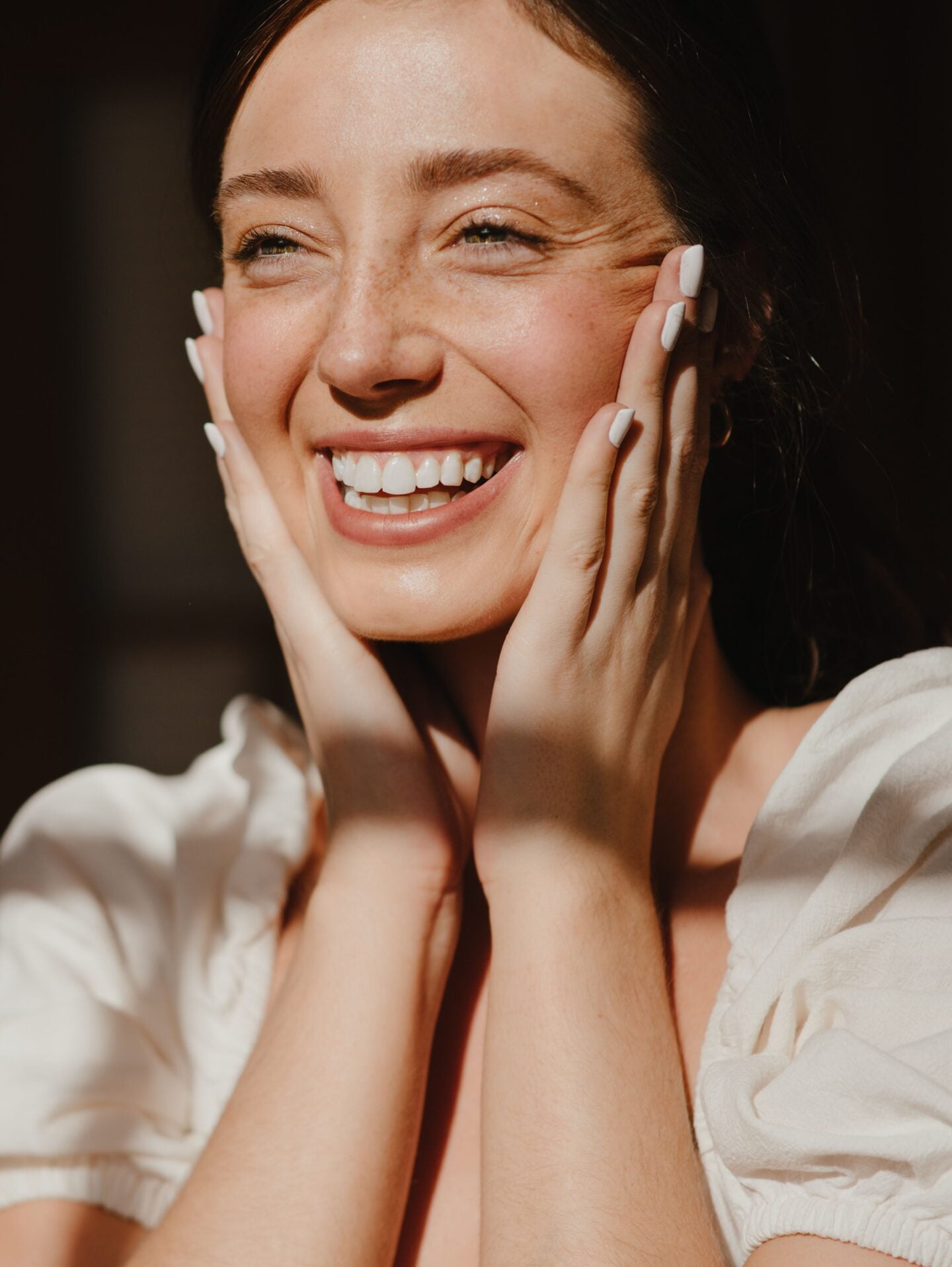 Close-up portrait of a smiling woman with dark hair, holding both hands against the sides of her face. She has white painted fingernails and is wearing a white puff-sleeve top. Warm sunlight falls across her face, hands and shoulders against a dark background.