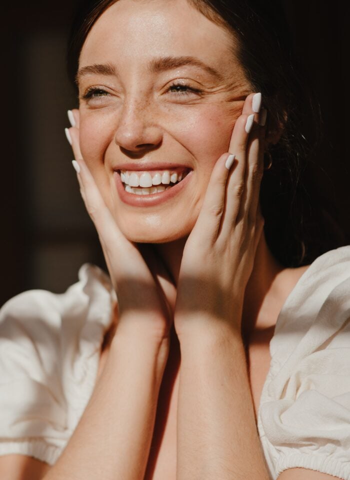 Close-up portrait of a smiling woman with dark hair, holding both hands against the sides of her face. She has white painted fingernails and is wearing a white puff-sleeve top. Warm sunlight falls across her face, hands and shoulders against a dark background.