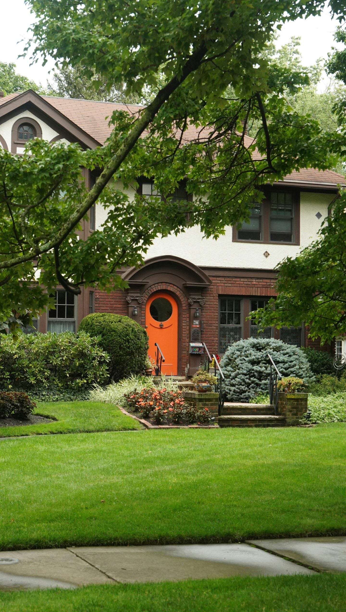 Front view of a two-story traditional home surrounded by mature leafy trees and lush landscaping, featuring a bright orange front door, red brick detailing, cream stucco walls, dark-framed windows, and steps leading to the entrance. The front garden includes neatly trimmed shrubs, flowering plants, evergreen bushes, and a large, healthy green lawn extending across the foreground beside the sidewalk.