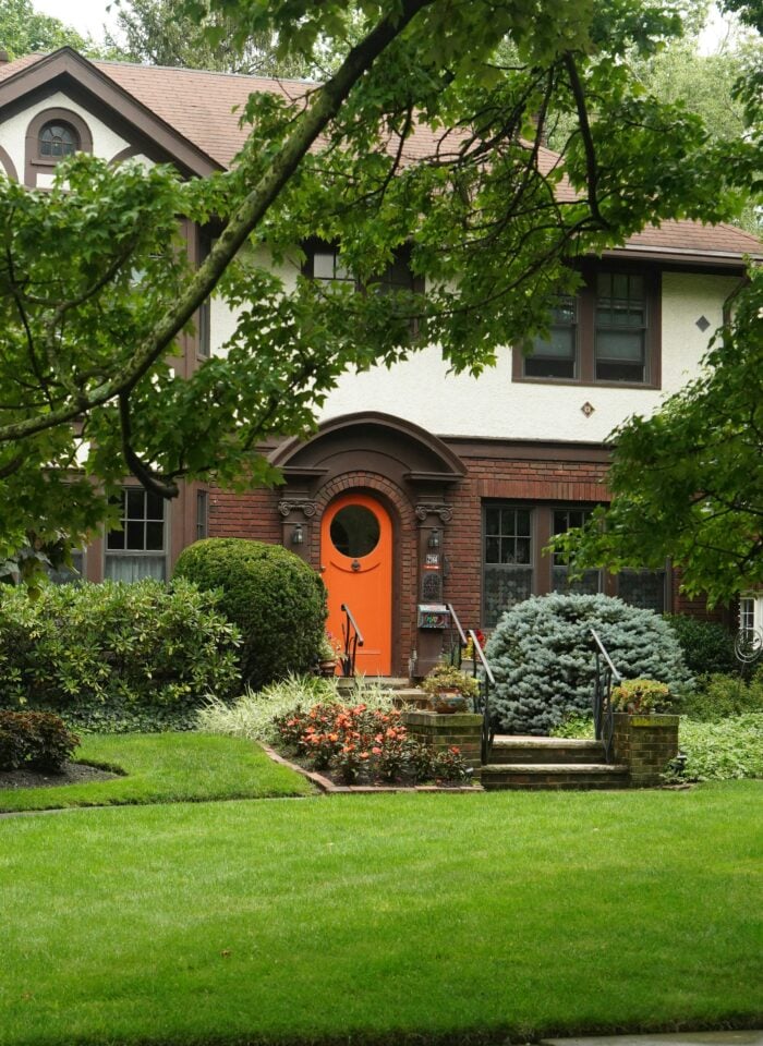 Front view of a two-story traditional home surrounded by mature leafy trees and lush landscaping, featuring a bright orange front door, red brick detailing, cream stucco walls, dark-framed windows, and steps leading to the entrance. The front garden includes neatly trimmed shrubs, flowering plants, evergreen bushes, and a large, healthy green lawn extending across the foreground beside the sidewalk.