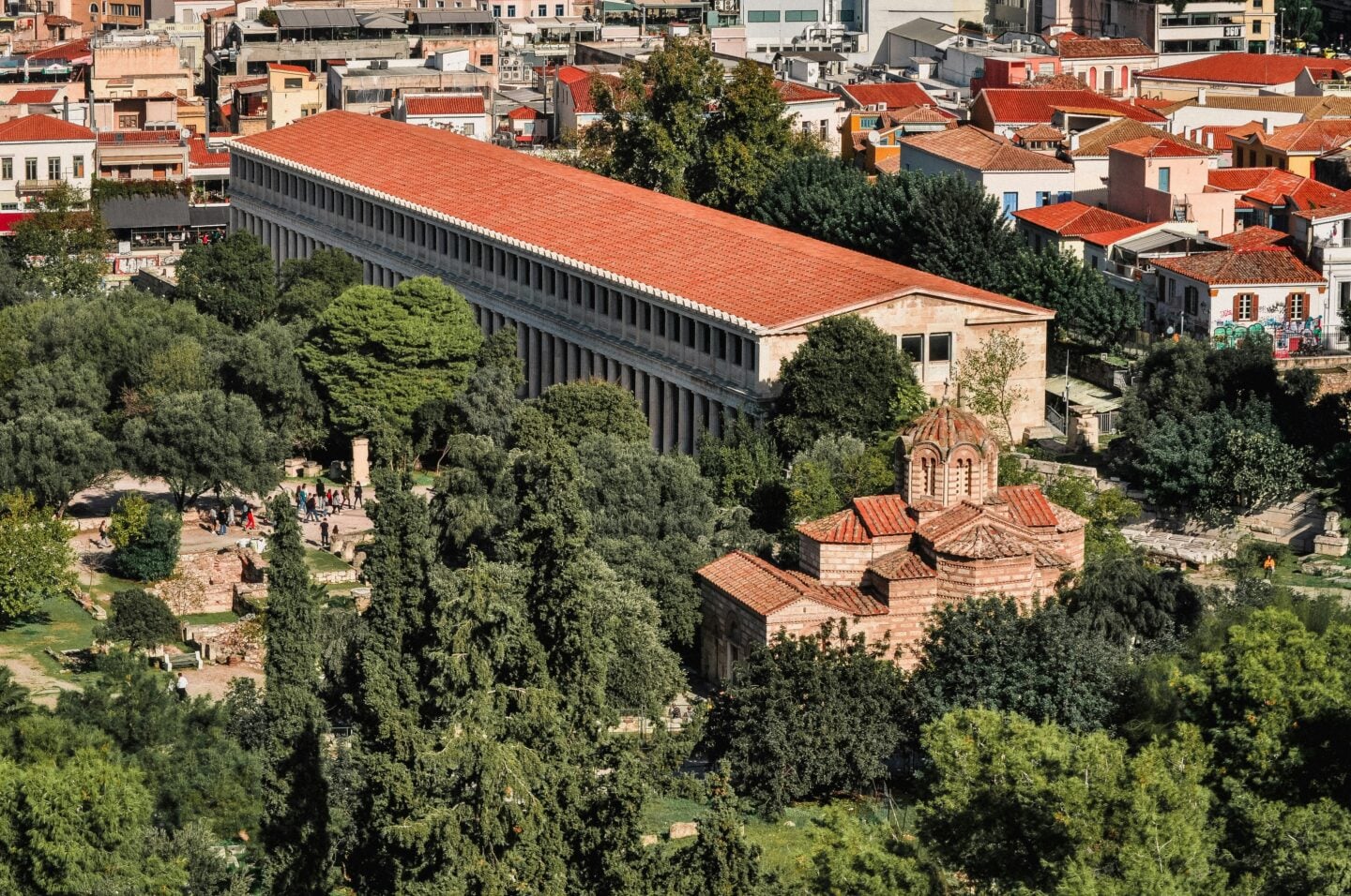 Stoa of Attalos: Elevated view of the Stoa of Attalos in Athens, showing its long colonnade and red-tiled roof amid trees, ancient ruins, and surrounding city buildings.
