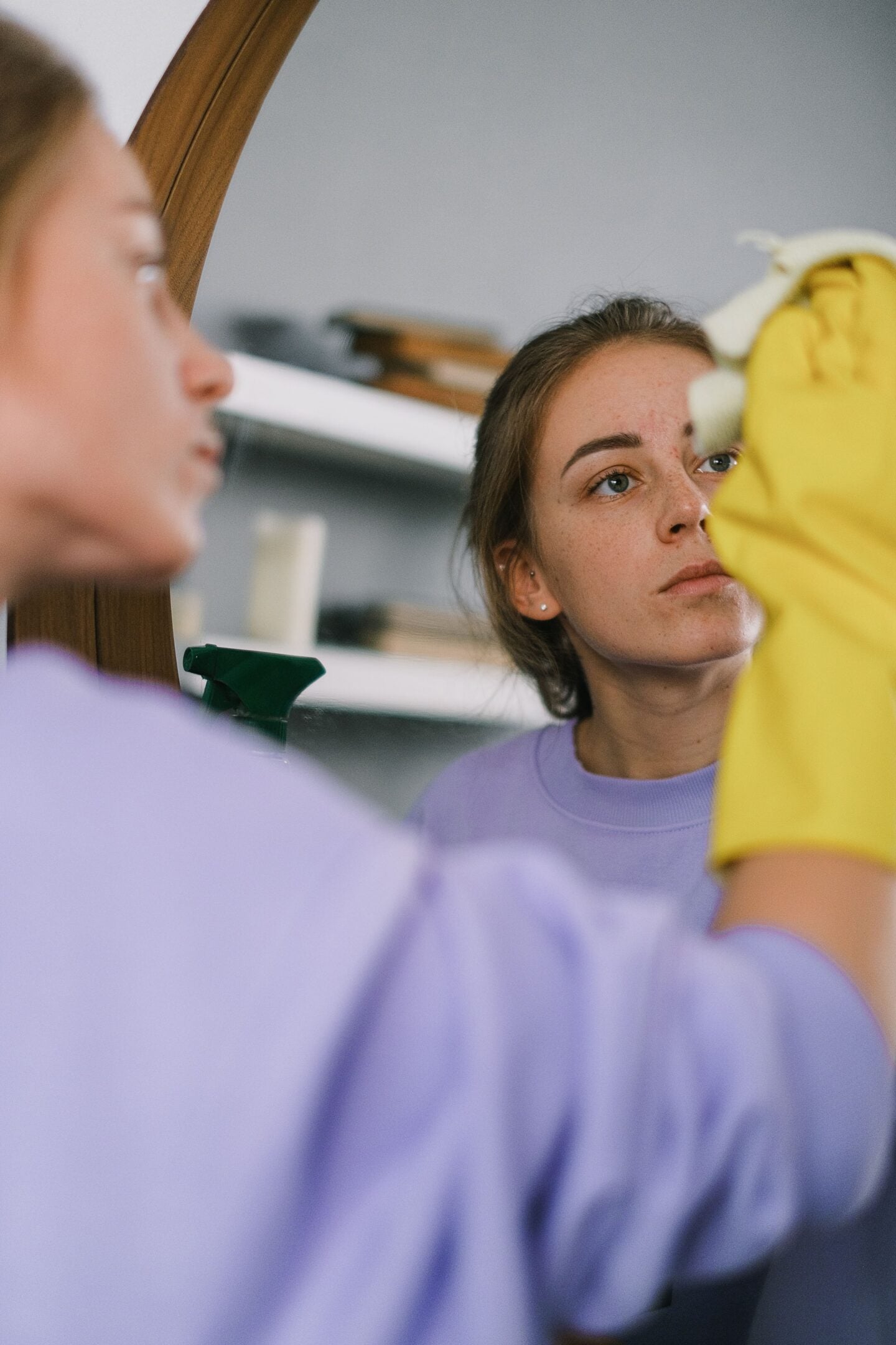 Woman wearing yellow rubber gloves cleaning a bathroom mirror with a cloth, with her reflection visible and a spray bottle nearby.