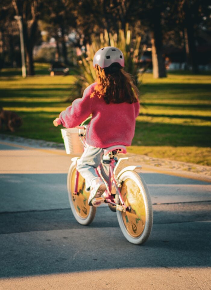 Child wearing a white helmet and pink fleece riding a pink bicycle with a front basket along a paved path through a sunlit park.