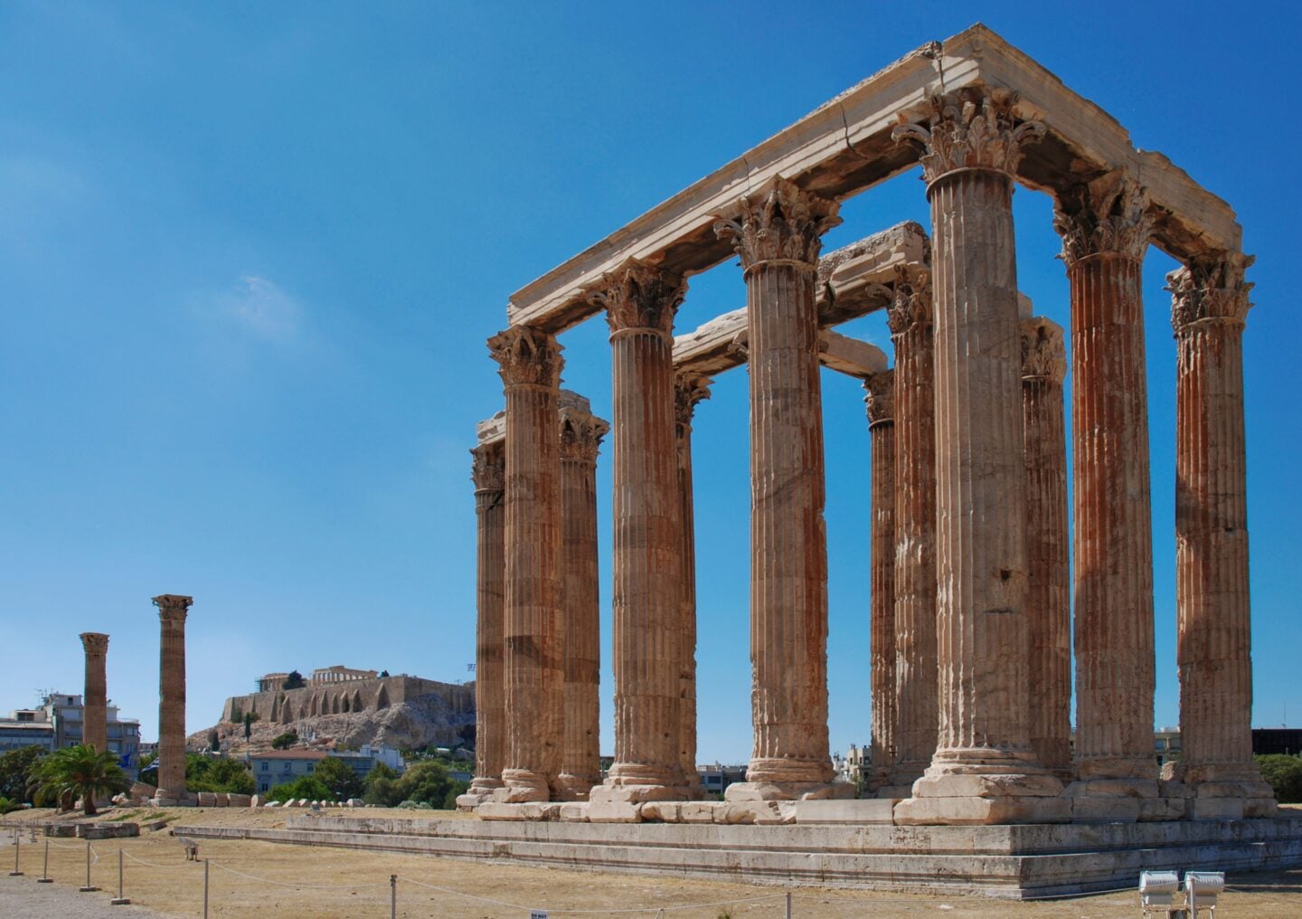 Temple of Olympian Zeus: Tall Corinthian columns of the Temple of Olympian Zeus in Athens, with the Acropolis visible in the distance beneath a clear blue sky.